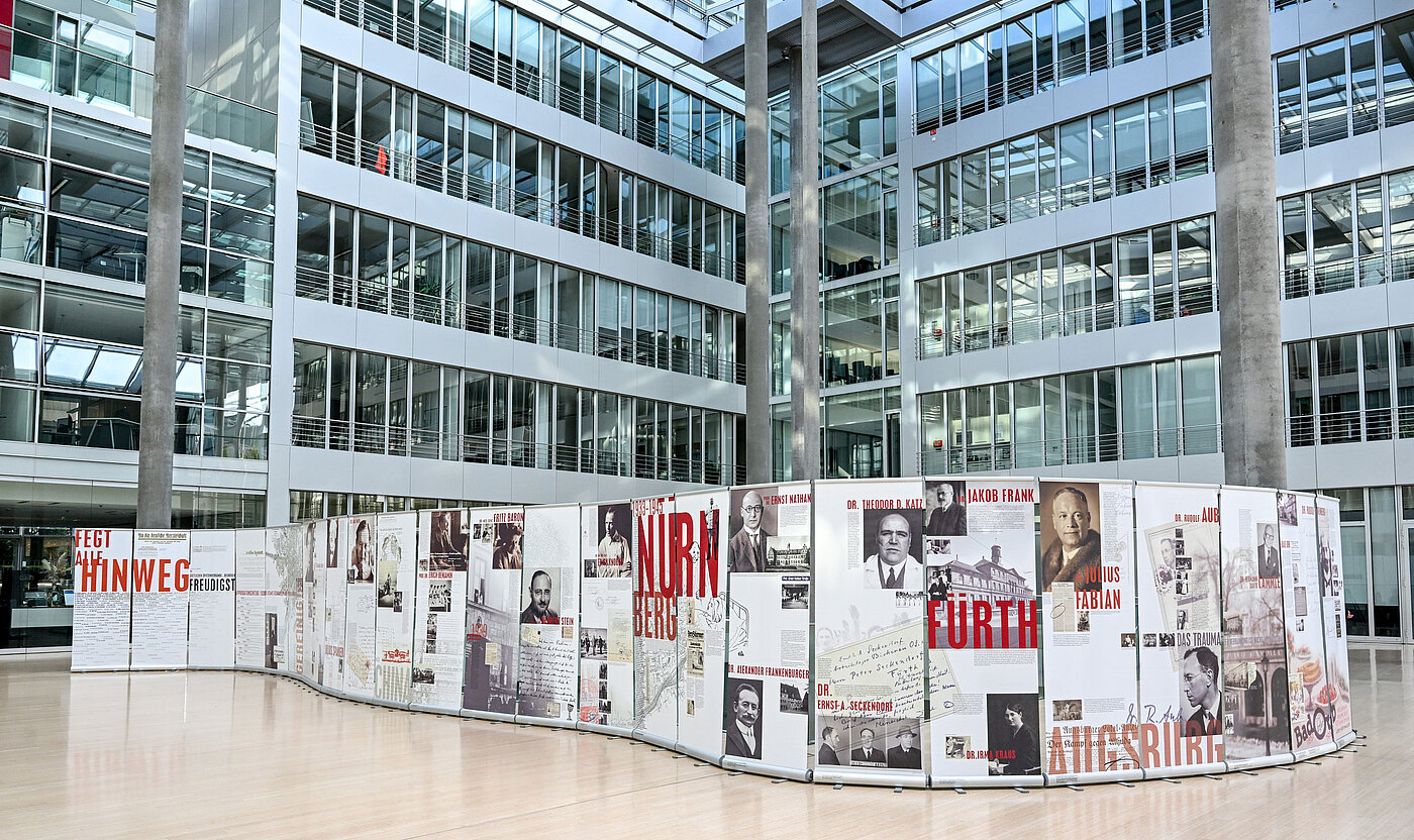 Das Bild zeigt eine Ausstellung mit Bildern und Texten auf mobilen Plakatwänden in einem Gebäudefoyer