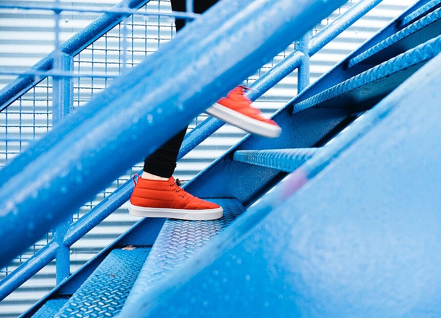 Das Bild zeigt eine Treppe, auf der jemand mit roten Sportschuhen nach oben steigt
