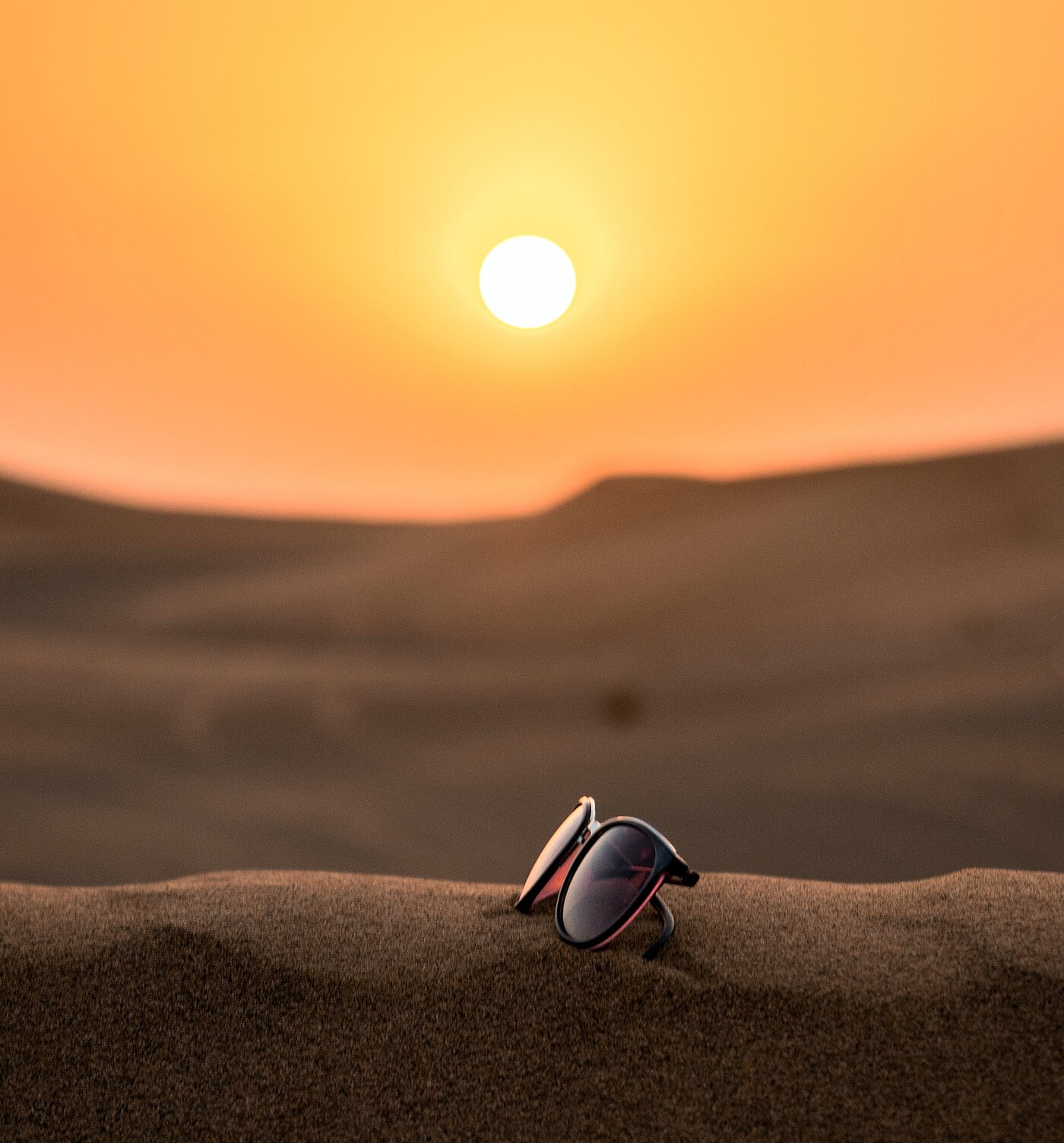 Das Bild zeigt eine Sonnenbrille, die vor untergehender Sonne auf einer Sanddüne liegt