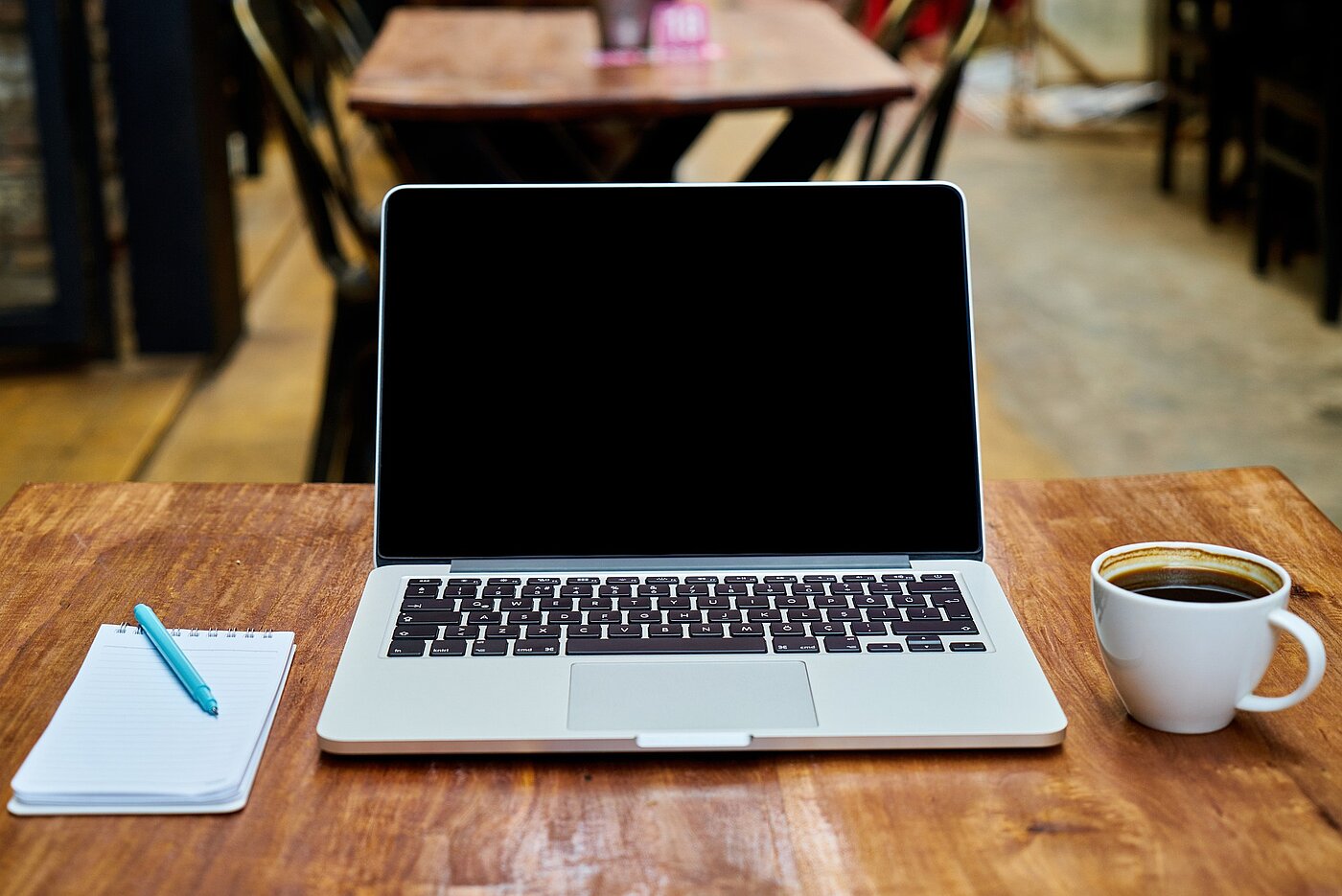 Foto eines aufgeklappten Laptops, daneben Kaffee und Block.