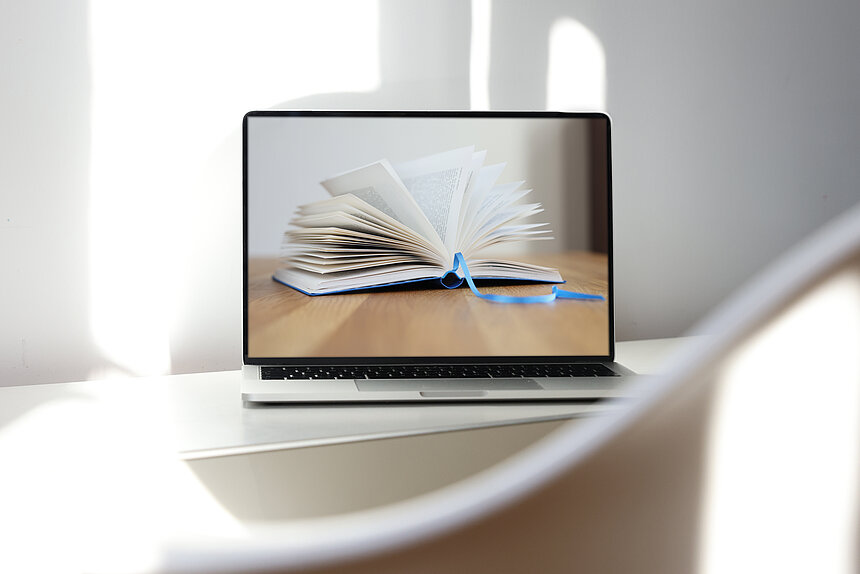 Das Bild zeigt einen Computer auf dessen Monitor ein aufgeschlagenes Buch zu sehen ist.