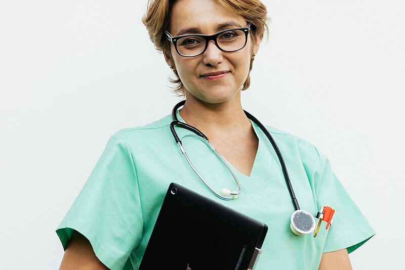 Das Bild zeigt eine Frau mit Stethoskop und Kladde in güner Krankenhauskleidung